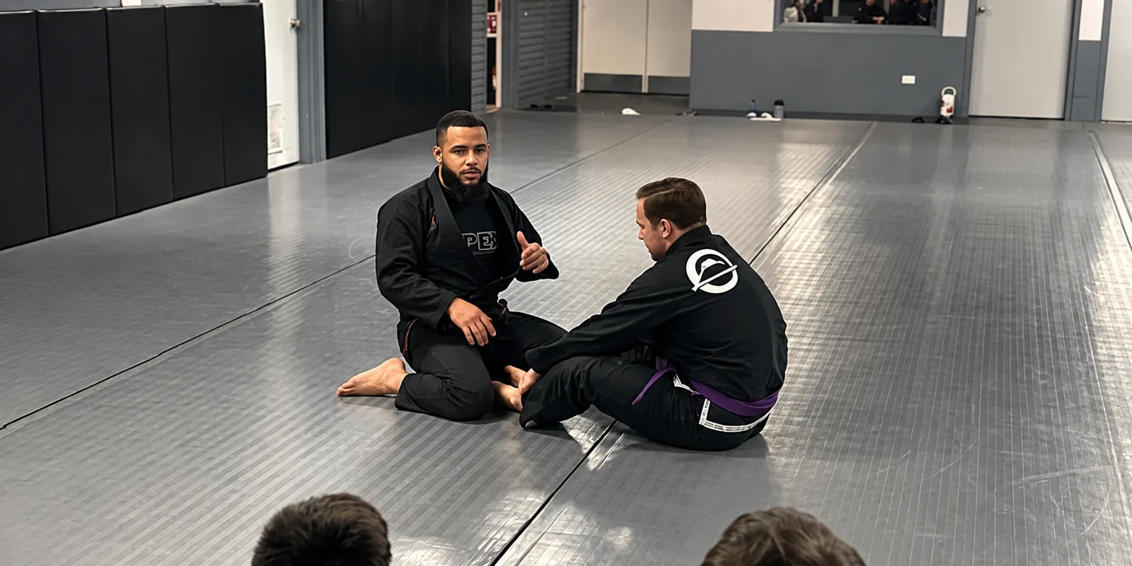 Coach Nick Spacek sitting on the mat explaining a technique to a purple belt while students watch