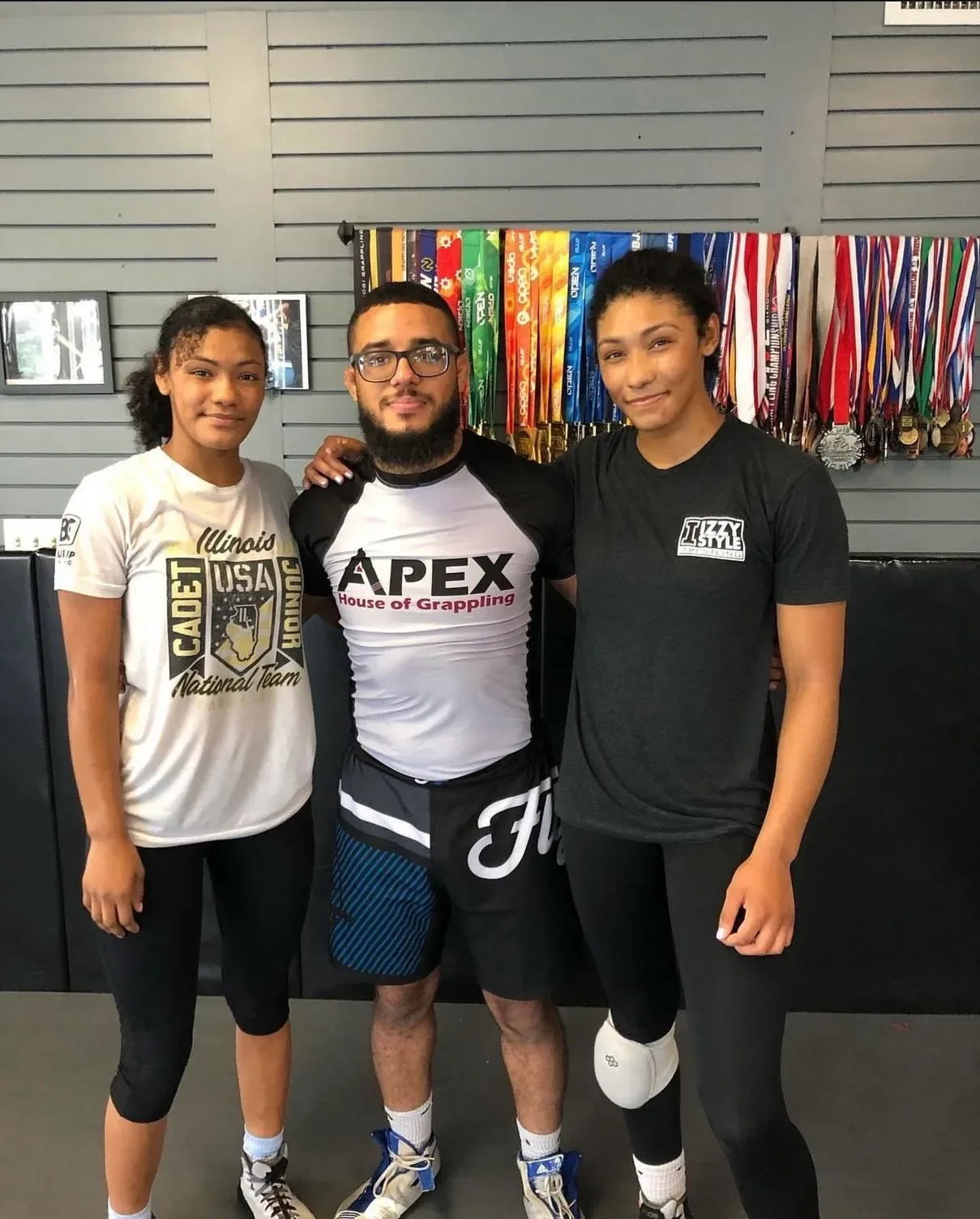 Coach Nick Spacek with Korina and Kennedy Blades in front of the Apex medal wall